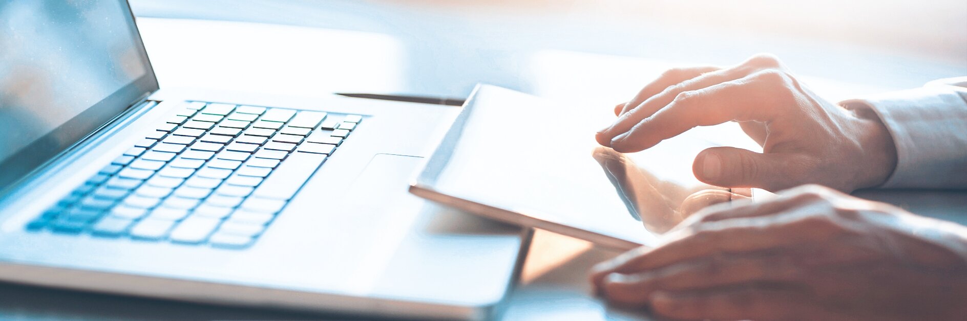 A person is using a laptop with their right hand on the keyboard in a bright, sunlit room. A smartphone and a closed book or notebook lie on the desk beside them. The background features a large window, creating soft lighting.