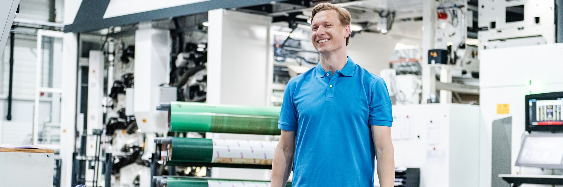 A man in a blue polo shirt and jeans is standing in a large industrial facility. Behind him, there is machinery and large rolls of tesa tape material. The setting appears to be a printing or manufacturing plant. (This text has been generated by AI)