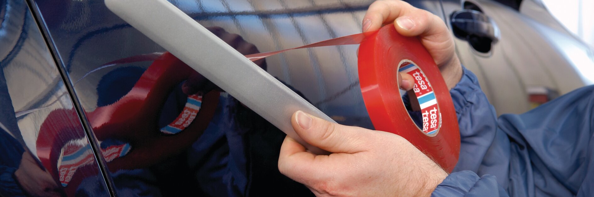 A person in a blue jacket is applying red tesa tape to the side of a dark-colored car. They are holding a tool with the tesa tape to ensure even application. The background shows a blurry interior of a garage or workshop.