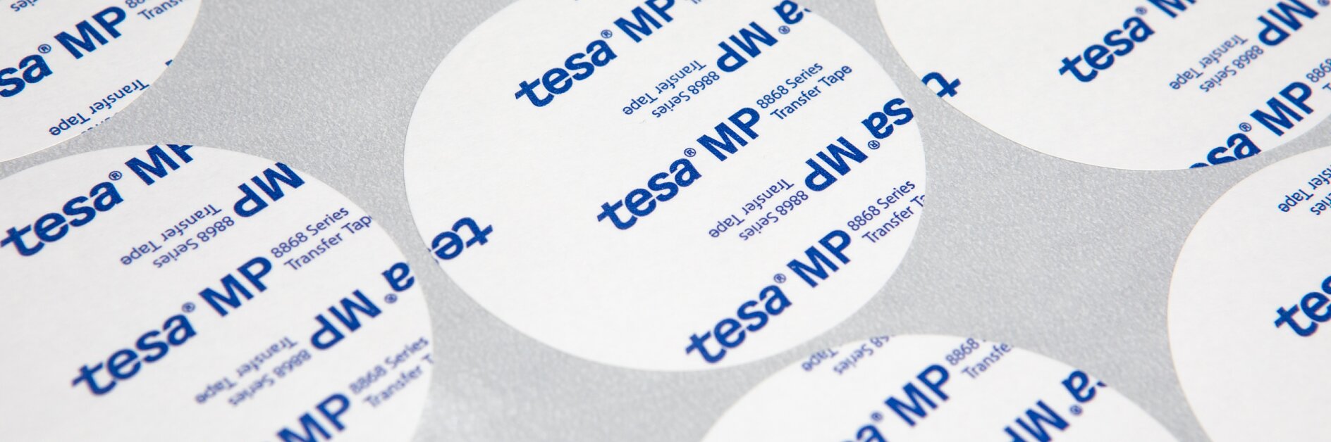 Circular adhesive patches on a surface. The patches have blue text printed with tesa MP and other text partially visible. They are arranged in a regular pattern, overlapping in places. The background is a plain, light-colored material. (This text has been generated by AI)
