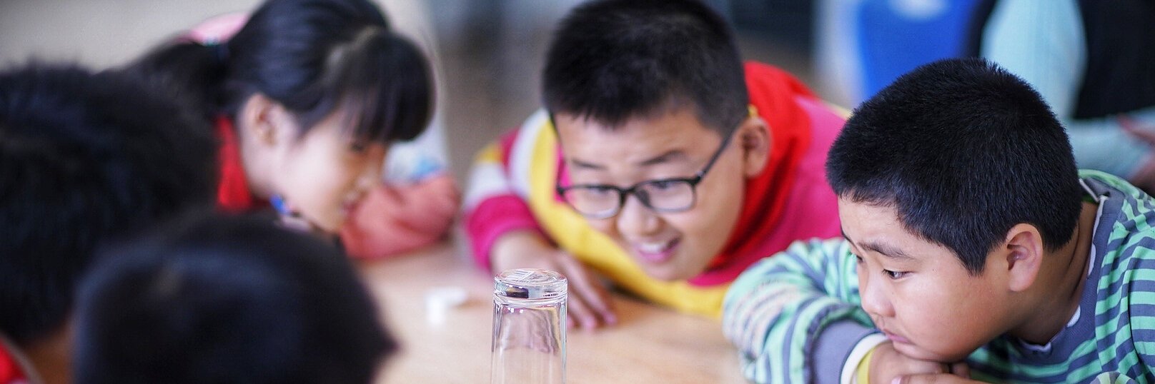 Children are gathered around a table, observing a science experiment. A lit candle is on a plate covered by an inverted glass, secured in place with tesa tape. One child is pointing at it, while the others watch attentively, suggesting they are curious about the experiment. (This text has been generated by AI)