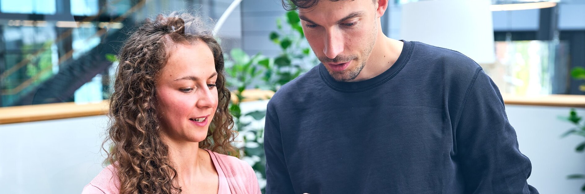 Two people are standing indoors, looking at a tablet. The woman on the left has curly hair and is wearing a pink sweater. The man on the right has short hair and is wearing a black sweatshirt. They appear to be discussing something on the screen, possibly related to tesa tape or its applications. (This text has been generated by AI)