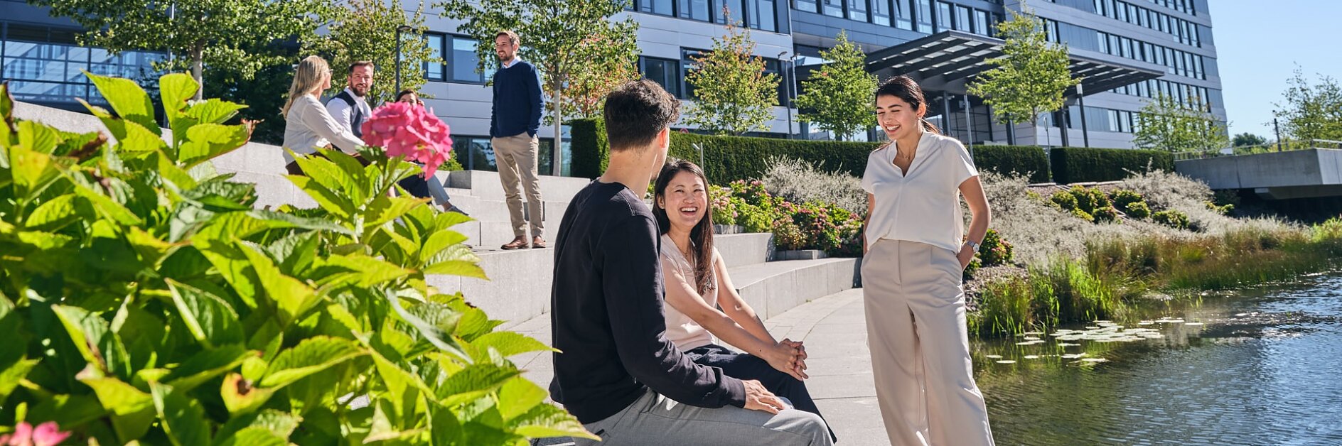People are sitting and chatting on steps by a small pond with greenery in front of a modern multi-story building. The sky is clear and sunny, and the building has a sign that is partially visible with tesa tape. (This text has been generated by AI)