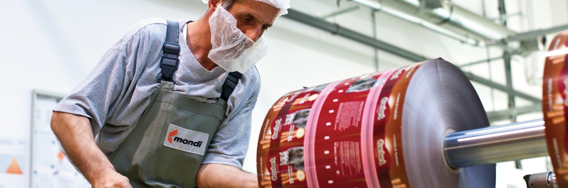 A worker wearing a hairnet and face mask inspects a large roll of printed packaging material in a factory setting. He holds a tool and is dressed in a gray shirt and green overalls. Machinery, tesa tape, and industrial elements are visible in the background. (This text has been generated by AI)