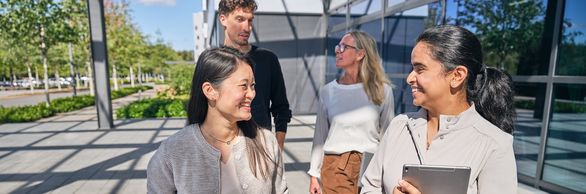 Four people walking and talking outside a modern building with large glass windows. Two women in the foreground, one holding a tesa tape tablet. Two individuals in the background, a man and a woman. Trees and blue sky visible in the background. (This text has been generated by AI)