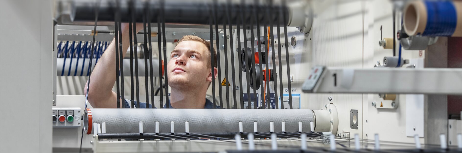 A person in a blue uniform works with machinery in a factory setting, adjusting black cables on a series of rollers and levers using tesa tape. The environment is industrial, with various mechanical components and controls visible.