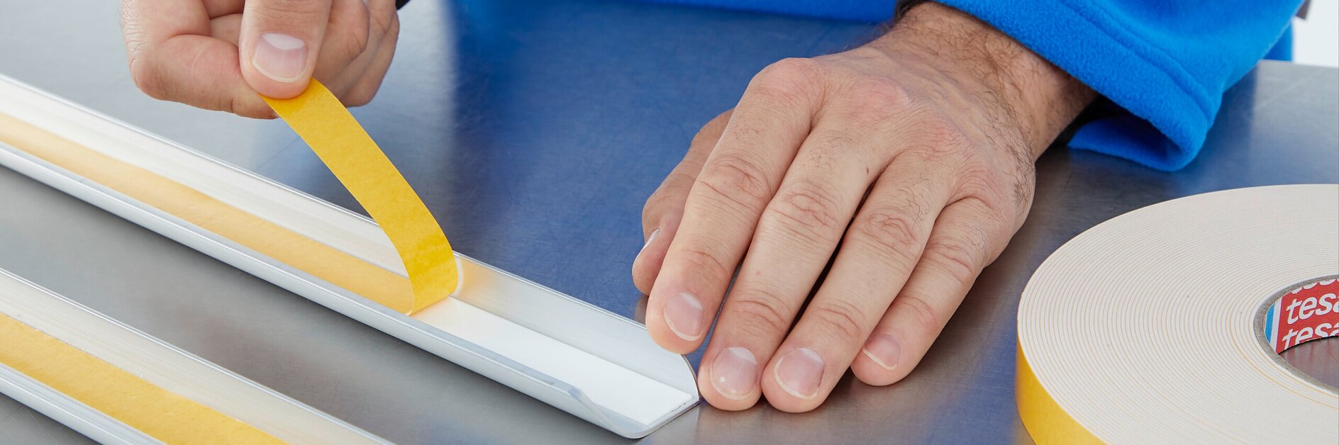 A person in a blue sleeve applies a strip of yellow double-sided tesa tape to a white rectangular object on a metallic surface. A roll of similar tesa tape is visible on the right side of the image.