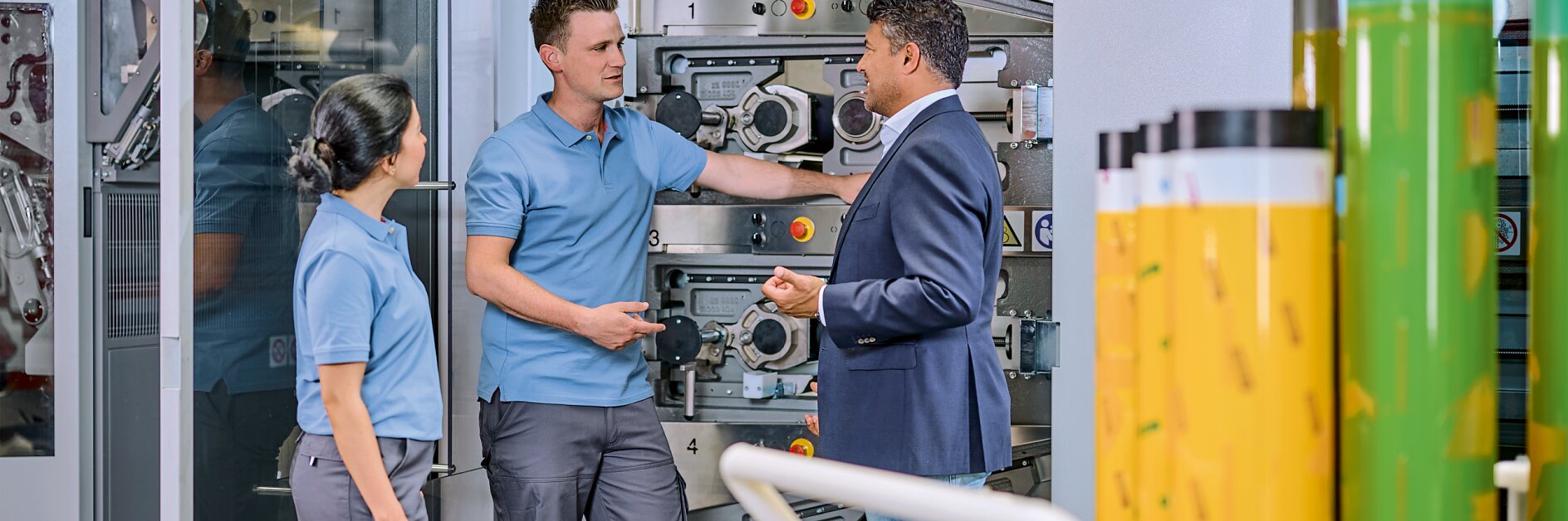 Three people stand in front of industrial machinery. Two individuals wear blue work uniforms, and one person in a blue blazer and jeans gestures while speaking. Rolls of tesa tape are visible in the foreground. (This text has been generated by AI)