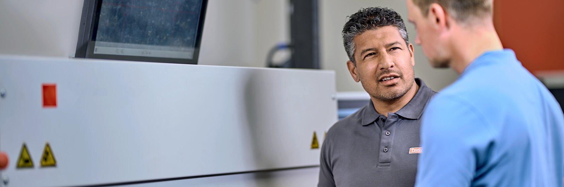 Two men are standing in front of a large industrial machine, discussing something. One man is holding a roll of tesa tape. A computer monitor is mounted on top of the machine. They are in a well-lit workspace. (This text has been generated by AI)
