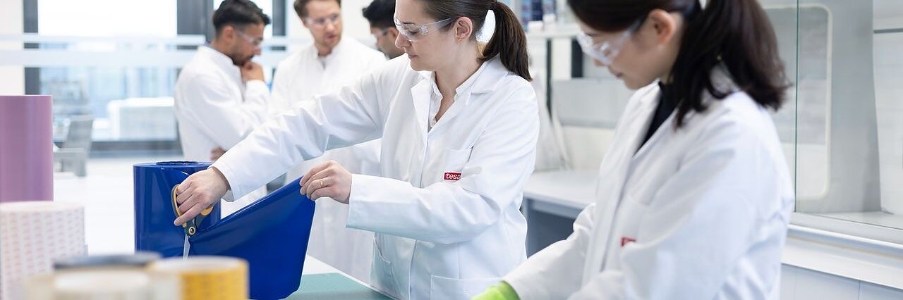 Several people in lab coats and safety glasses work with large rolls of material and tesa tape on tables in a bright laboratory, with shelves and lab equipment visible in the background. (This text has been generated by AI)